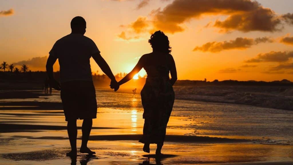 A silhouetted couple holding hands while walking on a beach at sunset.