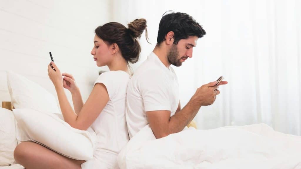A couple sitting back-to-back in bed, both looking at their phones.