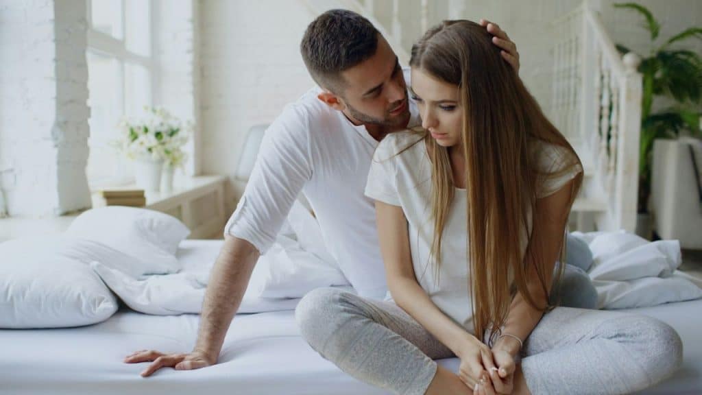 A man comforting a long-haired, sad-looking woman on a bed in a bright room.