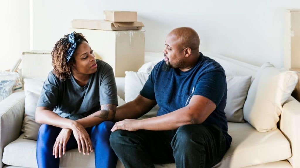 A man and a woman sitting on a couch talking, surrounded by moving boxes.