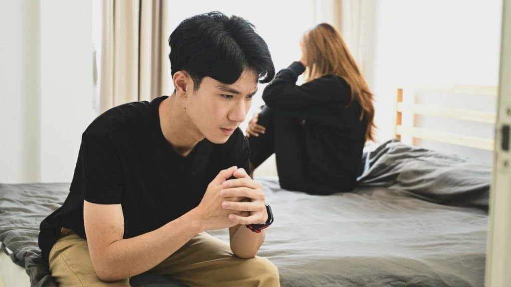 A pensive man sitting on a bed, with a woman crying in the background.