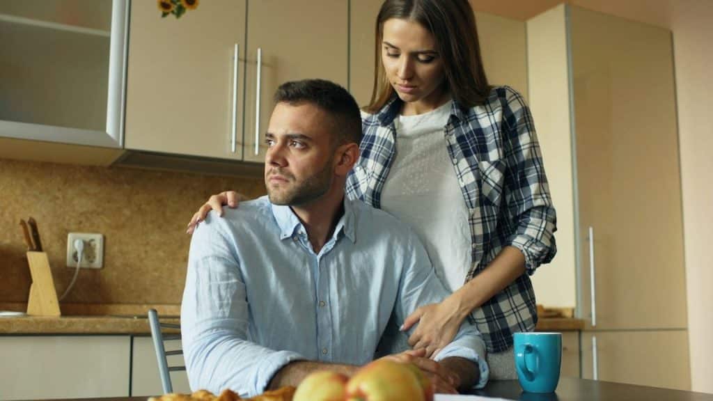 A woman in a plaid shirt comforting a serious-looking man seated at a table.