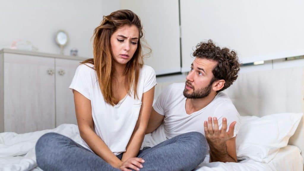 A worried-looking woman and a man talking, both sitting on a bed.