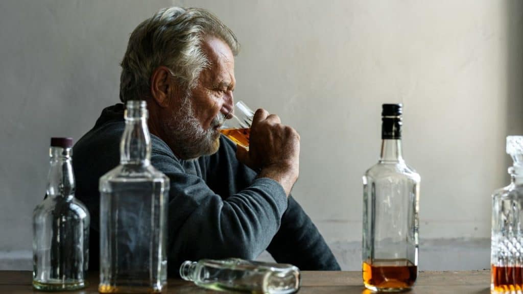 Senior man drinking from a glass, surrounded by several liquor bottles on a table.