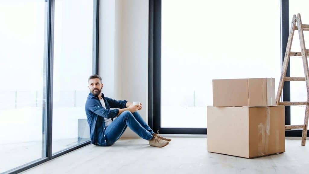 A man sitting on the floor next to large windows, surrounded by boxes and a ladder.