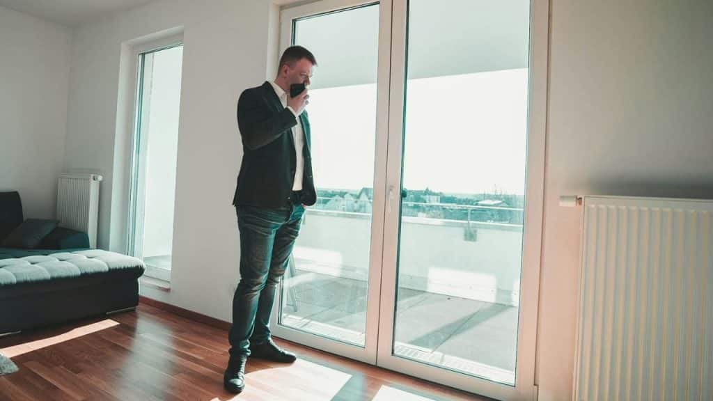 A man in a blazer and jeans standing by a glass door, looking out, holding a phone.