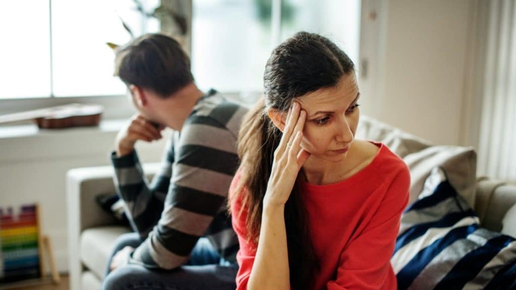 A woman in a red top with her hand on her head, looking upset, with a man beside her.
