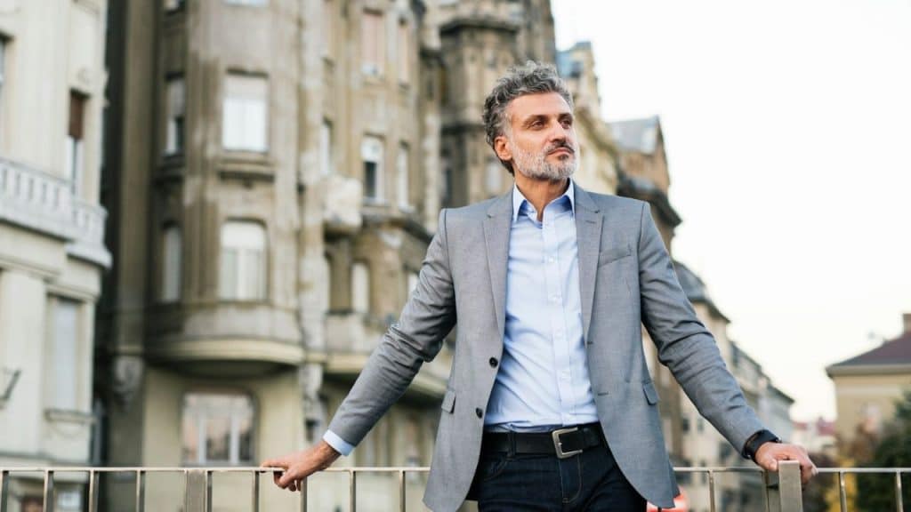 A distinguished, middle-aged man in a suit jacket leaning on a railing in an urban setting.