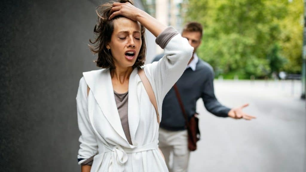 A distressed woman holding her head while a gesticulating man walks behind her outdoors.
