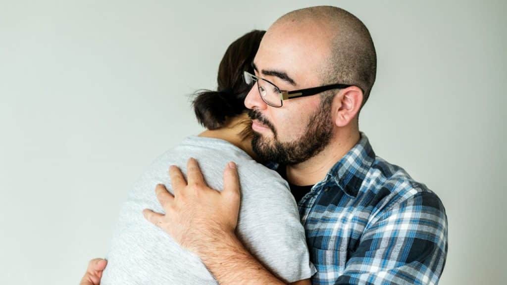 A man with a beard and glasses hugging another person tightly.