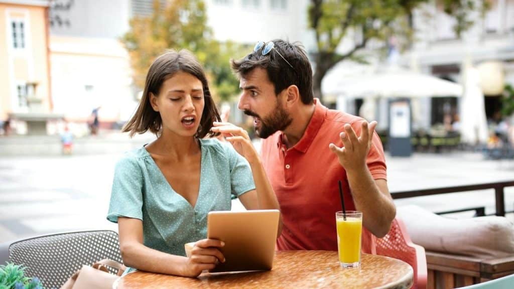 A man gesticulating and shouting at a woman holding a tablet outdoors at a cafe.