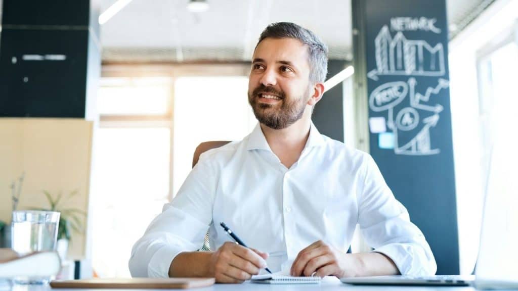 A smiling, bearded man in a white shirt writing at a desk indoors.