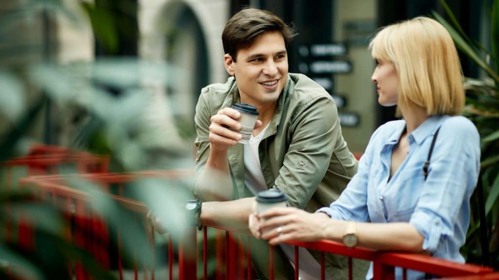 A smiling man and a woman talking outdoors while holding coffee cups.