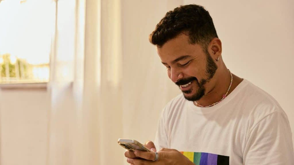A smiling, bearded man in a white shirt looking down at his phone.