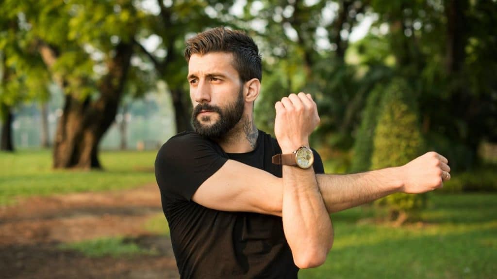 A bearded man in a black shirt stretching his arm outdoors in a park.