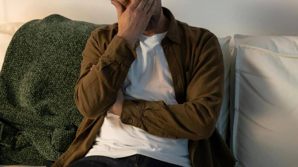 A man sitting on a couch covering his face with one hand.
