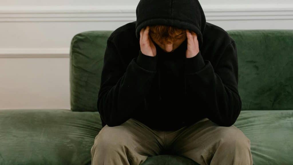 A person in a hoodie sitting on a couch with their head in their hands.