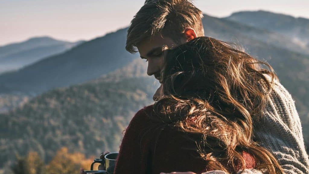 A couple sits together on a mountain overlooking scenic forested hills.