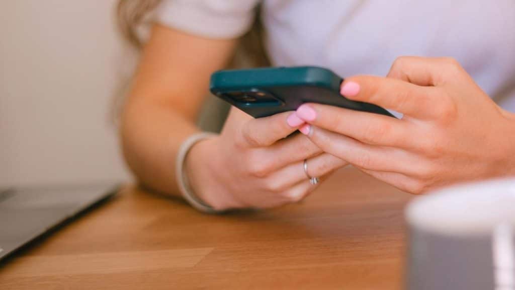 A person using a smartphone with pink-painted nails at a wooden table.