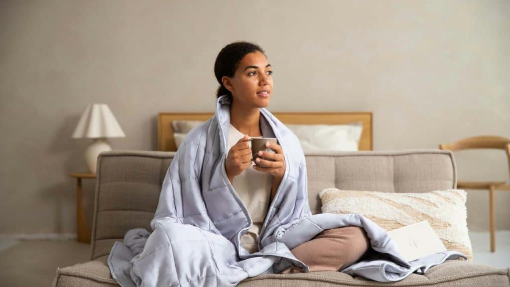 A woman wrapped in a blanket sitting on a sofa and holding a cup.
