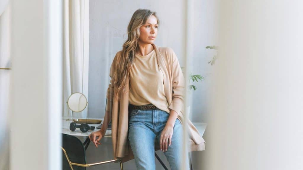 A woman in casual clothes sitting on a desk and looking thoughtfully to the side.