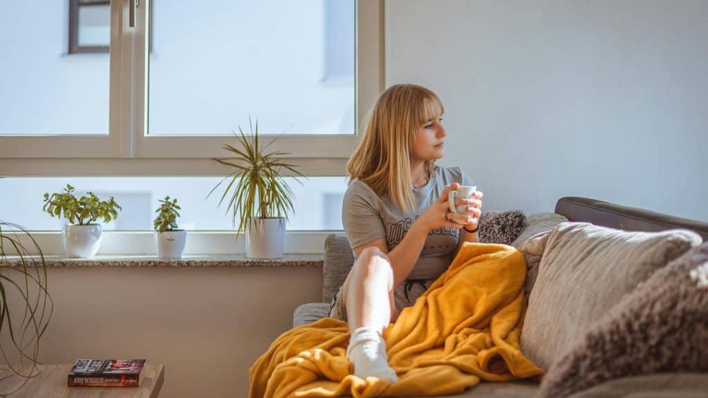 A woman sitting on a sofa with a yellow blanket, holding a mug and looking out the window.