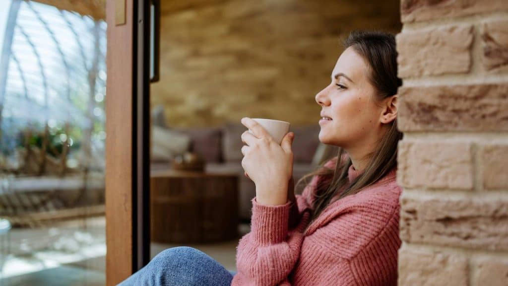 A woman sitting by a window holding a cup and smiling thoughtfully.