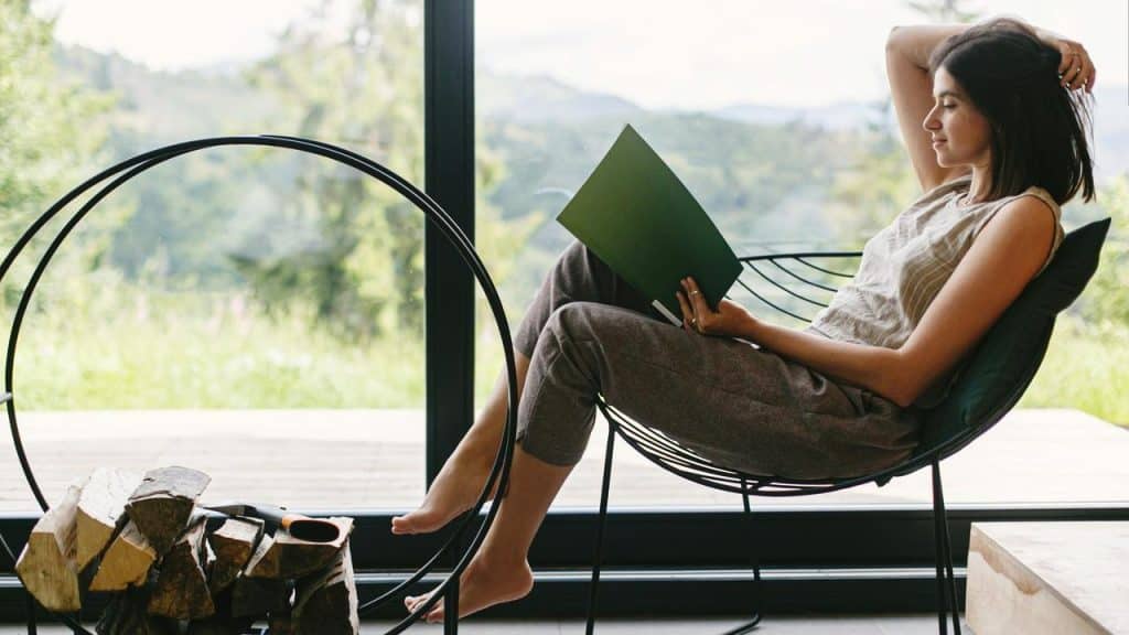 A woman sitting in a chair by a large window reading a book.