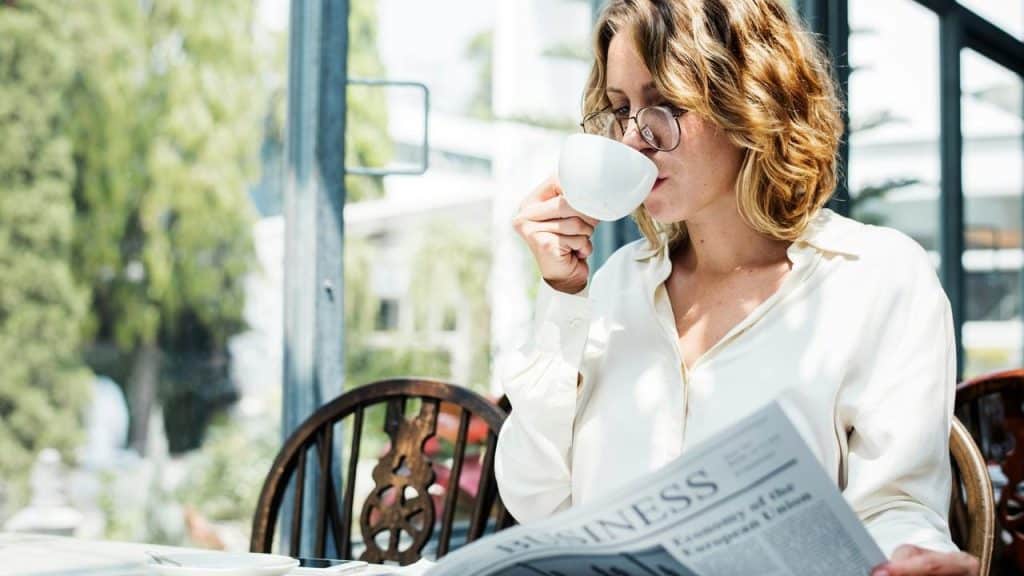 A woman wearing glasses drinking coffee while reading a newspaper.