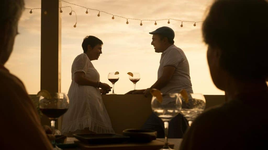 A couple talks by a sunset terrace with drinks and string lights in the background.