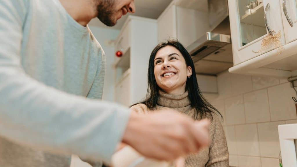 A couple smiles and talks while cooking together in a kitchen.