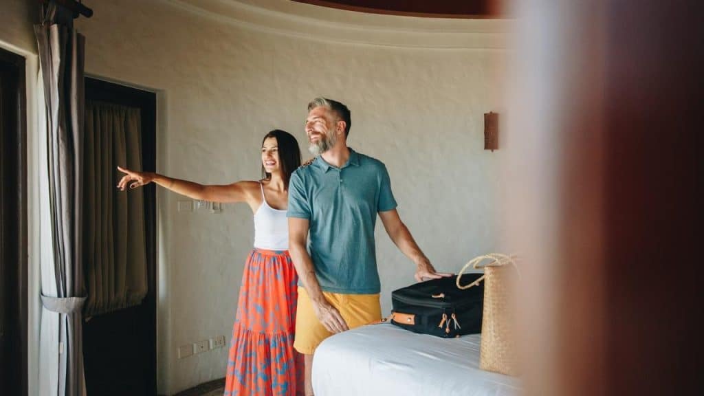 A couple stands in a bright hotel room, smiling and looking out the window.
