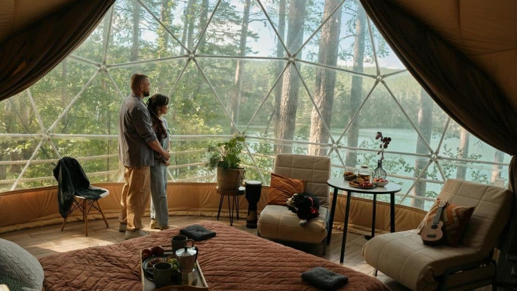 A couple stands inside a cozy dome cabin, looking out at a forest and lake view.