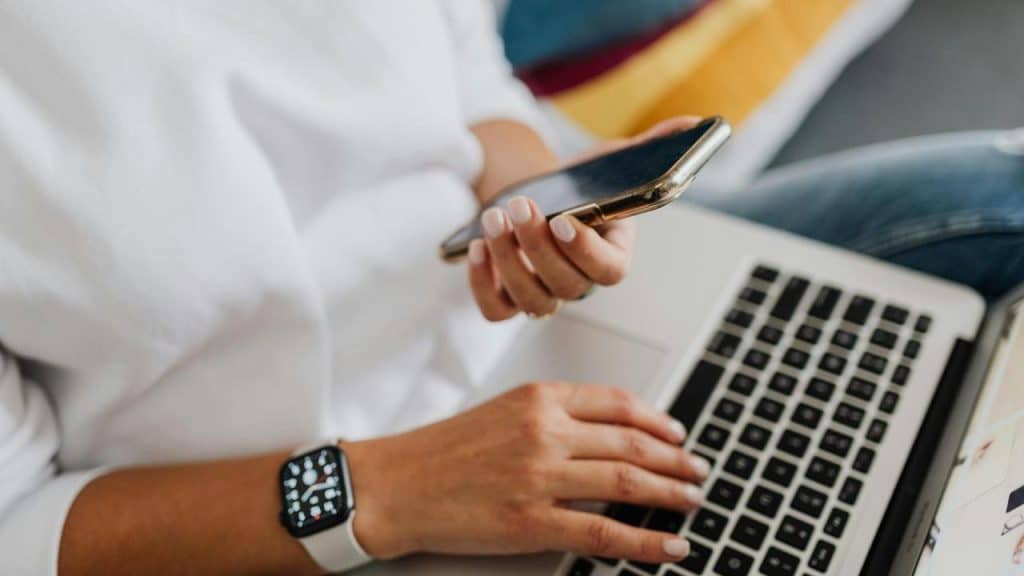 A person uses a laptop while holding a smartphone and wearing a smartwatch.