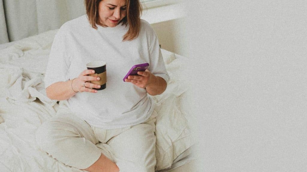 A woman sits on a bed holding a coffee cup, looking at her phone with concern.