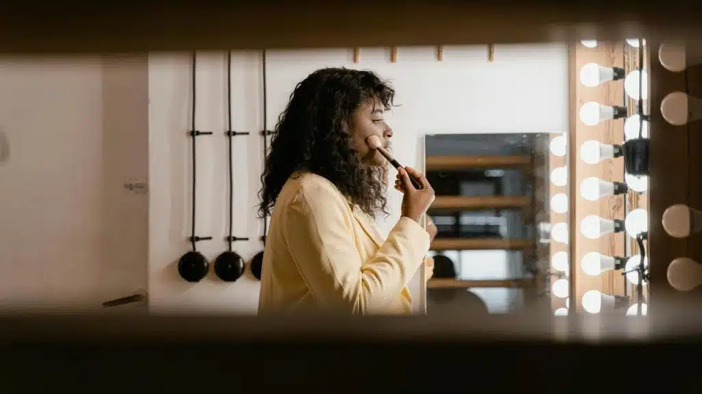 A woman applies makeup in front of a mirror surrounded by bright lights.