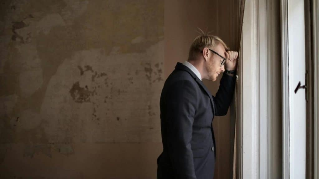 A man in a suit leans on a window, appearing deep in thought.