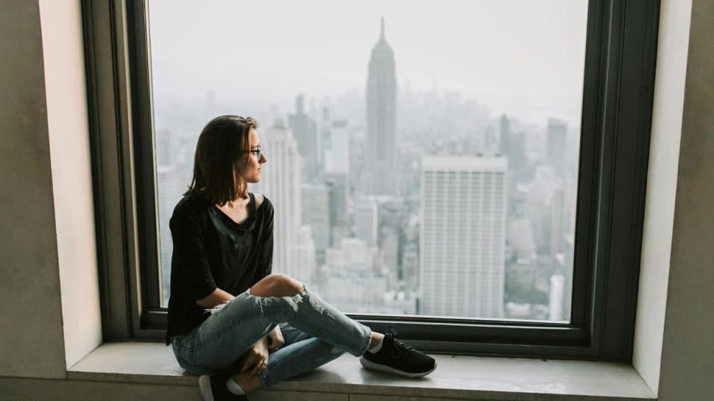 A woman sits by a large window, gazing out at a city skyline with tall buildings in the distance.