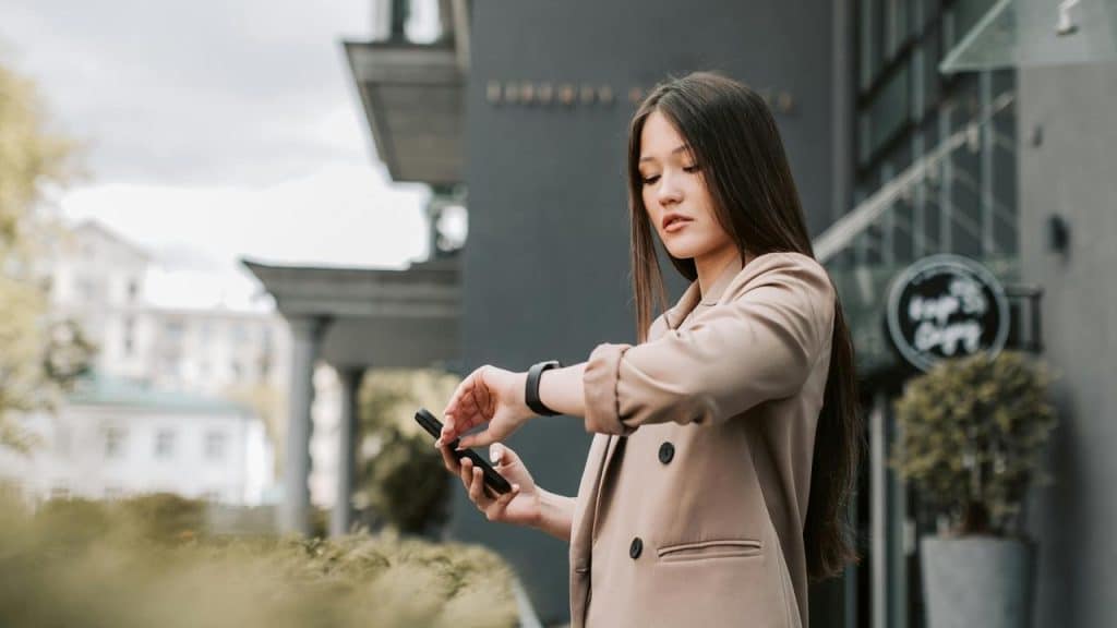 A woman stands outside holding a phone and checking the time on her smartwatch.