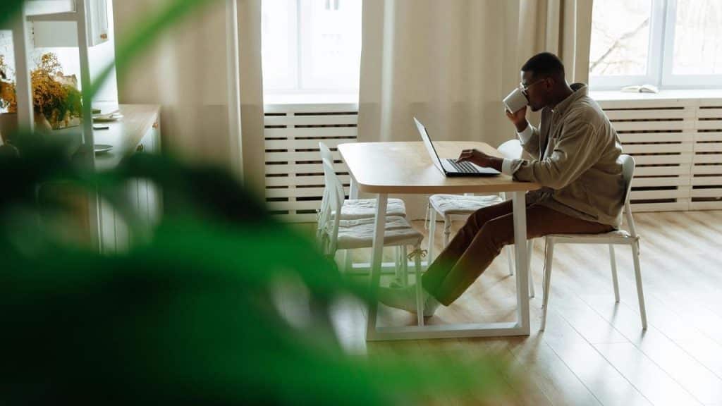 A man sits at a table working on a laptop while drinking from a mug.