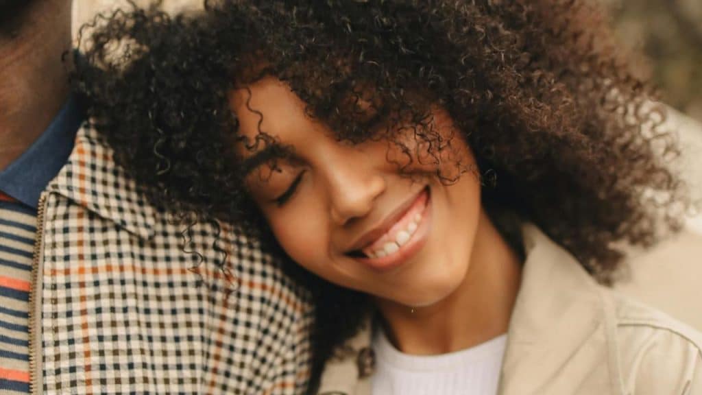 A smiling woman with curly hair rests her head on someone's shoulder.
