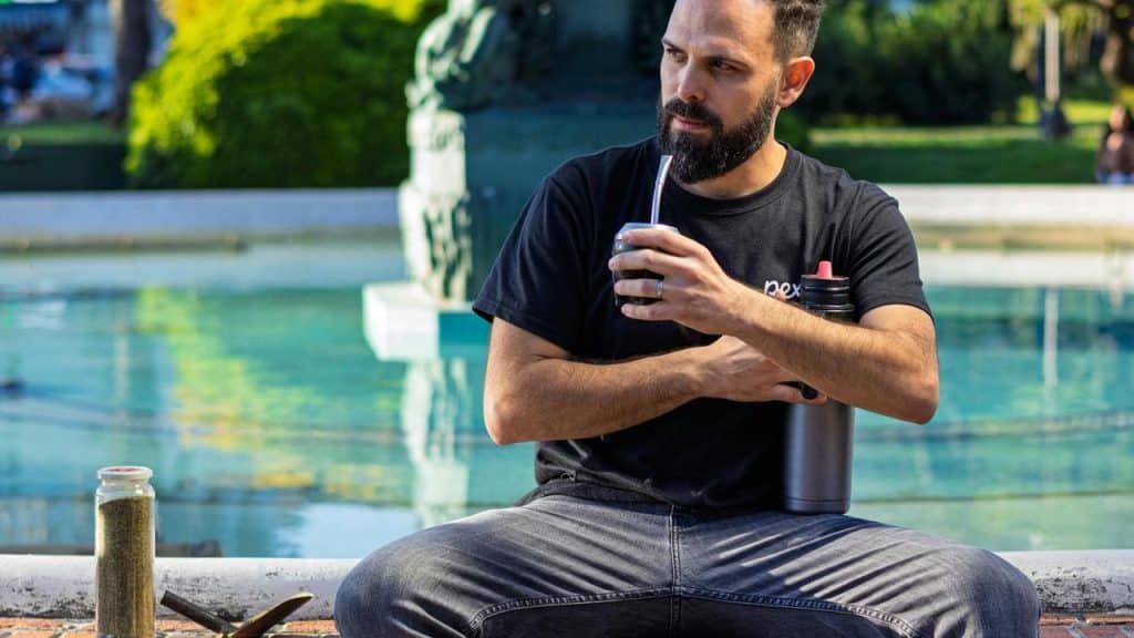 A man sitting by a fountain is drinking from a metal cup with a straw.