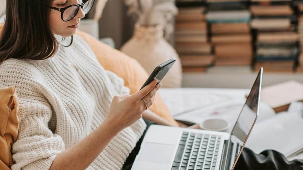 A woman wearing glasses looks at her phone while sitting with a laptop.