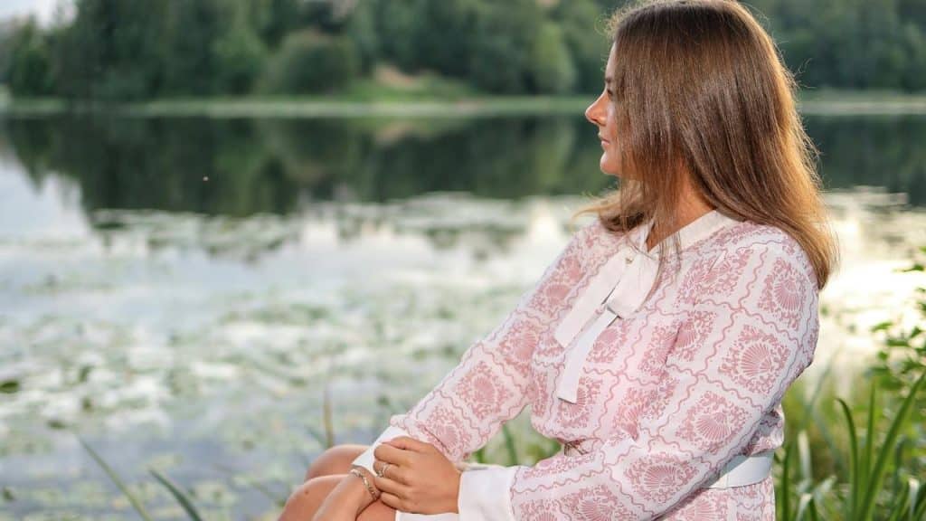 A woman in a pink patterned dress sits by a lake, looking at the water.