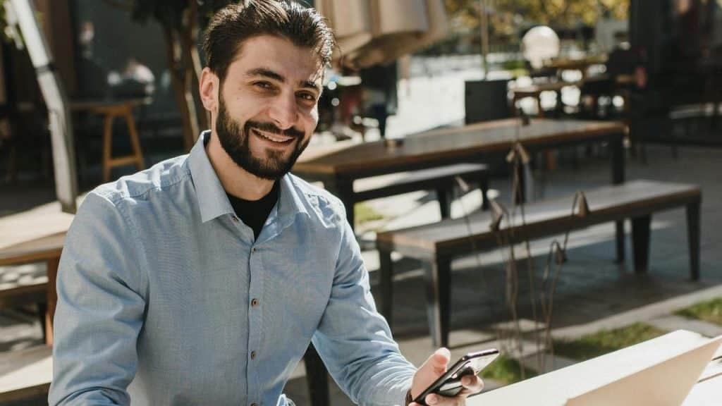 A man with a beard and blue shirt smiles while holding a smartphone outdoors.