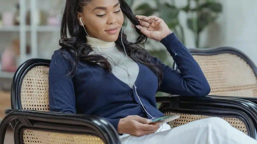 A woman sits in a chair listening to music with earphones.