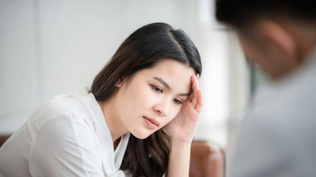 A woman is seated, holding her forehead with a hand, looking stressed.