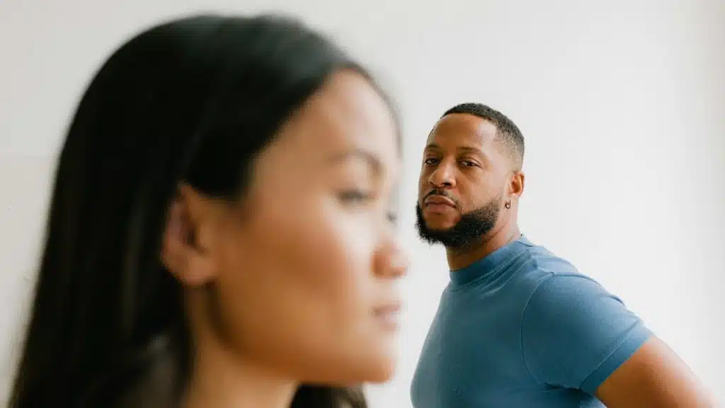 A focused shot of a bearded man in a blue shirt looking intently at the camera, with a woman's blurred profile in the foreground.