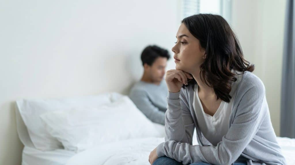 A young woman sits on the edge of a bed while a man sits away from her in the background.