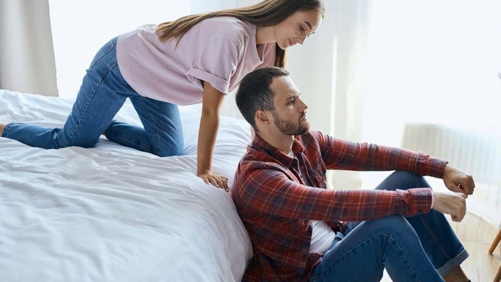 A man sits on the floor with his back against the bed, looking upset or thoughtful, while a woman leans over the bed toward him.